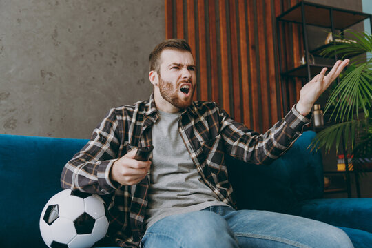 Young sad unhappy man fan in brown shirt hold remote controller spread hand cheer up support football team sit on blue sofa with soccer ball rest watch tv indoors room gray wall Sport leisure concept