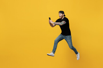 Fototapeta na wymiar Full body side view young bearded tattooed man 20s he wearing casual black t-shirt cap jump high run fast hold in hand use mobile cell phone isolated on plain yellow wall background studio portrait