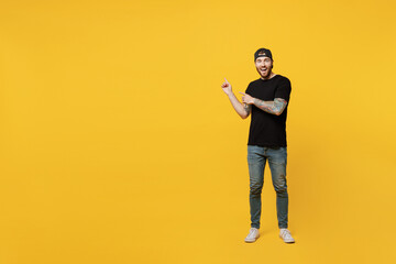 Fototapeta na wymiar Full body young amazed bearded tattooed man 20s he wears casual black t-shirt cap indicate point index fingers aside on workspace area mock up isolated on plain yellow wall background studio portrait