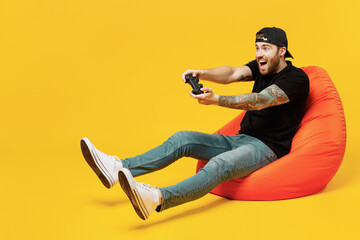 Full body young bearded tattooed man 20s he wears casual black t-shirt cap sit in bag chair hold in...