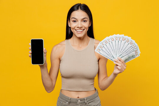 Young latin woman 30s she wear basic beige tank shirt holding fan of cash money in dollar banknotes use mobile cell phone with blank screen workspace area isolated on plain yellow backround studio