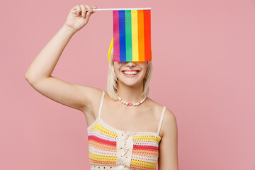 Young happy blond lesbian woman 20s she wear colorful knitted top yellow hat hold cover eyes with striped flag isolated on plain pastel light pink background. People lgbtq lifestyle fashion concept.