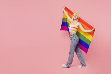 Full body young blond lesbian woman she wear colorful knitted top yellow hat hold striped flag look camera walk isolated on plain pastel light pink background People lgbtq lifestyle fashion concept.