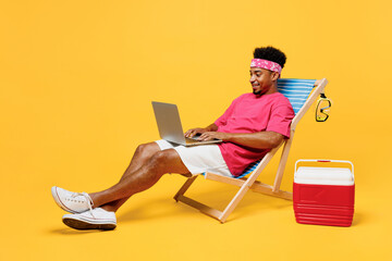 Full body side view smiling fun young man 20s he wearing pink t-shirt bandana near hotel pool hold use work on laptop pc computer isolated on plain yellow background. Summer vacation sea rest concept.