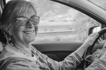 Black and white portrait of attractive happy senior woman with eyeglasses driving the car with hands over the steering wheel