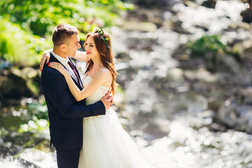 Young groom in a blue suit hugs in a park with green plants with a beautiful curly-haired bride in a long white dress. Against the background of the river.
