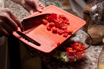 Naklejka na ściany i meble Chopped pieces of red bell pepper are poured into bowl with salad from board with knife. Cooking. Selective focus. Image for articles about food, dinners, peppers.