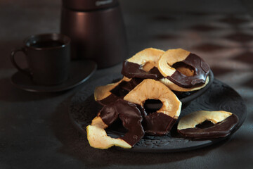 Apple chips with chocolate on a ceramic plate. Dried apples. Dried fruits from apples with chocolate. The geyser coffee maker is out of focus. A light snack.Vertical photo. Gobo mask.