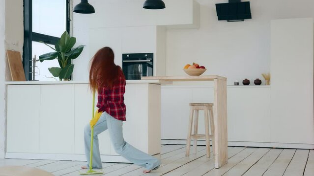 Active happy caucasian housewife amusing young redhead woman with long hair energetic dancer moving to music cleaning apartment flat washing floor with mop dancing in living room housekeeping chores