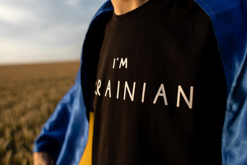 a man stands in a wheat field with the flag of Ukraine draped over his shoulders, wearing a black...