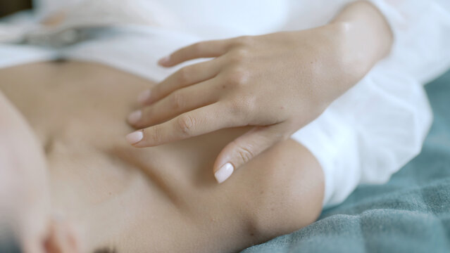 Close up of a young woman touching gently her collarbones while lying on bed in white dress. Action. Tender female relaxing on bed.