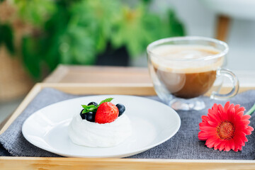 Pavlova dessert mini cake, cup of latte coffee and fresh gerbera flower on the wooden tray with gray napkin on the table. Coffee time. Breakfast for loved one. Take a break and relax. Selective focus