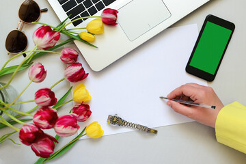 Top view of a desk where a woman's hand is writing on a piece of paper, next to it lies a laptop and a mobile phone, a bouquet of flowers, and space for text.