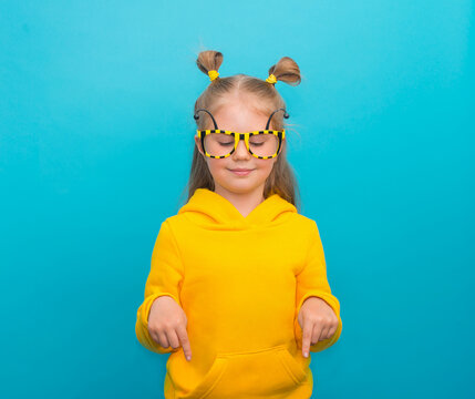 Funny smiling little girl in bee glasses and yellow hoodie on blue background  pointing down to empty copy space.