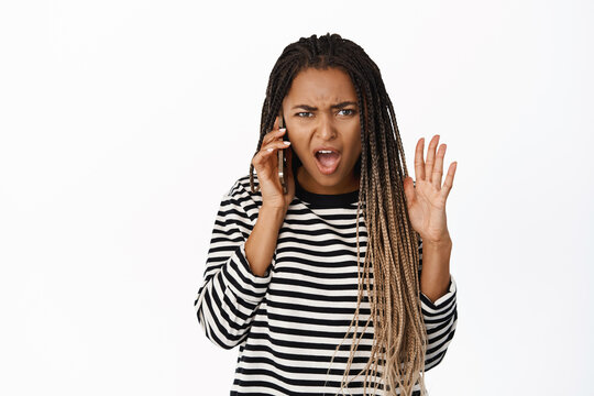 Shocked black woman answer phone call, talking on mobile with frustrated angry face, being insulted during conversation, white background