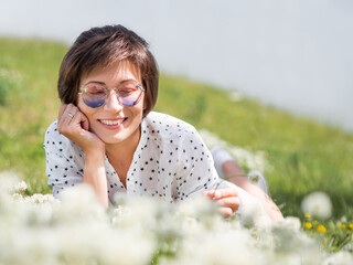 Woman in colorful sunglasses sniffs clover flowers on lawn in urban park. Nature in town. Relax outdoors after work. Summer vibes.