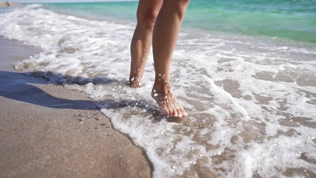 Beautiful Slim Female Legs Walk Barefoot along Sea Coast. Woman Tourist on Summer Vacation. Pretty Girl Walks at Seaside Surf. Unrecognizable Person. Splashes of Water and Foam in Super Slow Motion.