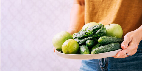 A woman holds in her hands fresh green vegetables fruits in large plate, light background copy space