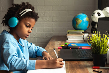 African-American girl doing homework,making notes,using headphones and laptop at home.Back to...