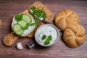 Spicy tzatziki with cucumber slices on fresh bun and chopped herb ingredients on cutting board.