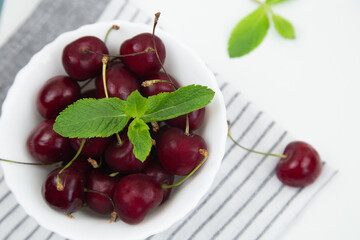 A white plate with a ripe burgundy cherry and a mint petal stands on a striped kitchen towel on a white background, top view. High quality photo