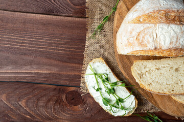 Close-up of a still life made of fresh crusty bread. A slice of bread smeared with cottage cheese and decorated with micro greenery. Linen napkin brown wooden background, flat lay, copy space