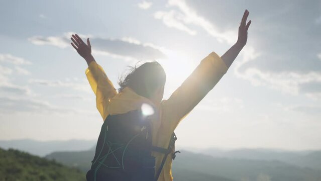 A happy girl in a yellow jacket raises her hands uphill on top of a mountain. Victory and freedom woman conquered the top of the mountain in a beautiful landscape at sunset. High quality 4k footage