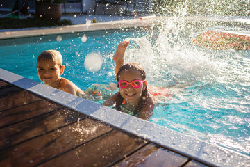 Cheerful children in googles laughing while playing in swimming pool at sunny day, refreshing at...