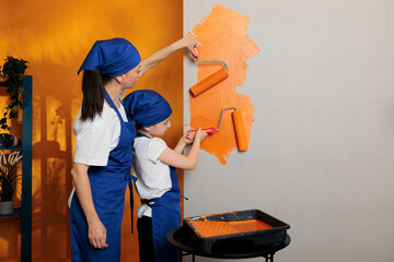 Young people painting walls with orange color paint and roller brush, doing housework renovation together. Mother with little kid using paintbrush and redecorating tools in apartment.