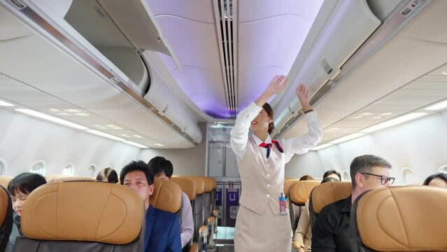 Asian woman flight attendant walking on airplane aisle closing luggage compartment over passenger seat before aircraft take off runway. Cabin crew checking safety and passenger security in the plane