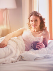 Young woman drinking cup of coffee or tea while lying in bed.