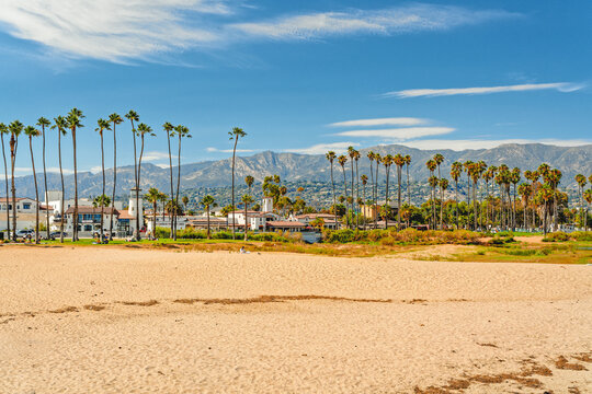 Wide sandy Santa Barbara beach with beautiful palm trees, city architecture in silhouette, mountains, and cloudy sky on background