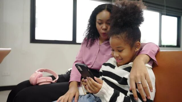 4K African family mother and little daughter using mobile phone together while waiting for flight boarding in airport terminal. Mom and kid go to travel destination by airplane on holiday vacation