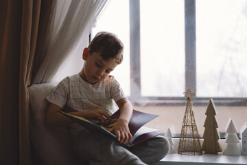 Cute boy is reading a book sitting on the windowsill near the window. Cozy home with decorative Christmas trees. Sweet home. Winter holidays lifestyle.