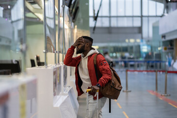 Upset unhappy African student guy standing with luggage and passport in airport terminal with frustrated facial expression, feeling stressed and uncertain about relocation to another country