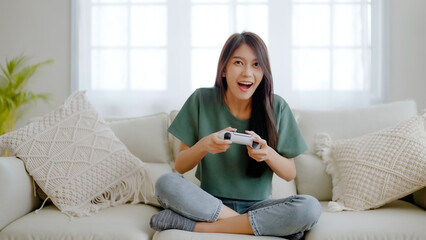 Beautiful young asian woman sitting on sofa and holding joystick playing online video game in...