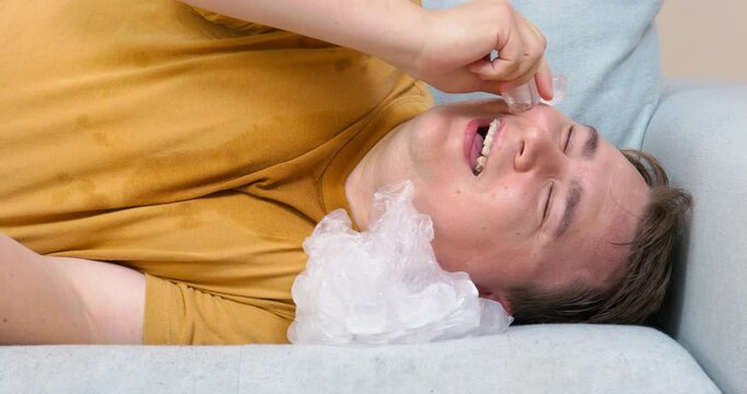 Young man is languishing from heat, so he lies on couch with ice pack, and puts one cube to face to cool off. Guy froze ice cubes with useful components and now does facial massage with their help