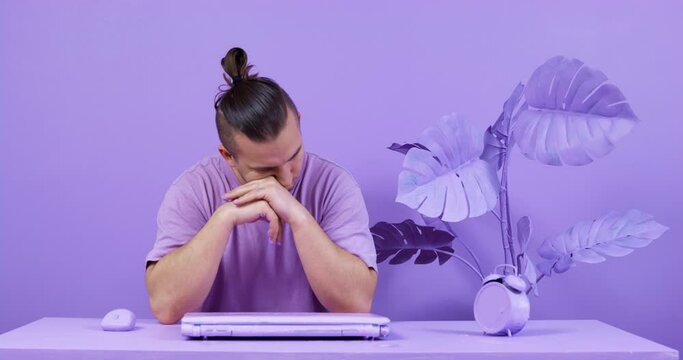 Young man is unable to complete difficult task and has no inspiration for work, so he sits in front of laptop, sighs heavily and shakes his head hopelessly. Everything is painted purple
