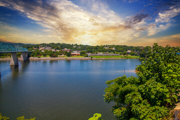 Fototapeta na wymiar a gorgeous summer landscape on the Tennessee River with the Walnut Street Bridge over the rippling blue water surrounded by lush green trees, grass and plants with powerful Clouds at sunset