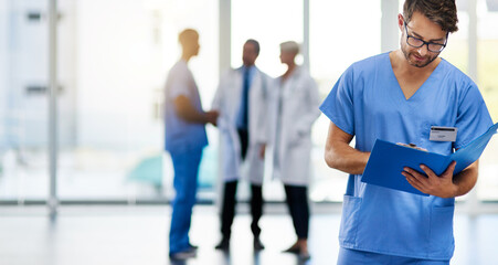 Young male doctor reading, holding and looking at patient health information at a hospital. Medical professional standing with doctors in the background. Healthcare worker working on a clinic file - Powered by Adobe