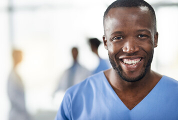 Happy, smiling and professional doctor in a hospital closeup portrait with blurred background. Confident black male medical healthcare worker with colleagues in the back. An African American surgeon.