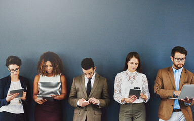 Group of businesspeople texting, typing and browsing online while waiting and standing in line on grey studio wall. Diverse team using tablet, laptop and phone and holding different wireless gadgets