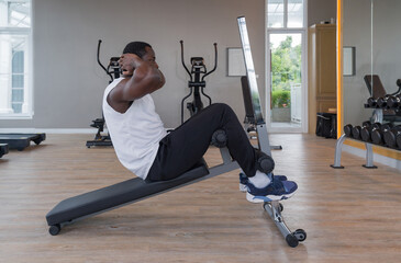 Young short curly black hair man with moustache and beard Sit-up on Multi-Function Bench. There are cardio machines and dumbbell set with rack in the gym.
