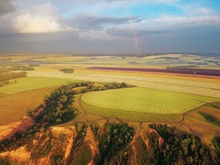 From a height, a view of high hills with fields of grass and groves of birches. Erosion of clay hills. Rainbow illuminated by the evening sun