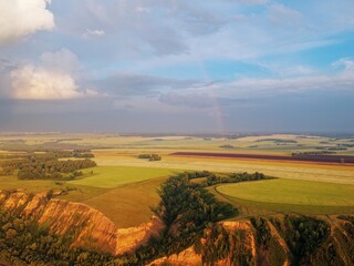 From a height, a view of high hills with fields of grass and groves of birches. Erosion of clay hills. Rainbow illuminated by the evening sun