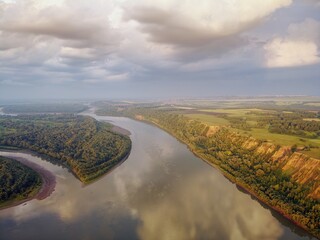 Riverbed under the mountain and groves of trees on the shore. Evening light of the sun. Clouds in the sky and reflection in the water. River Ob