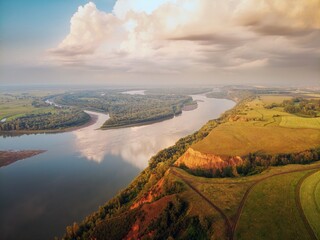 In large clouds, the light of the setting sun and the warm evening light flood the trees on the hill, the clay mountain and the reflection of the clouds in the water. river Ob.
