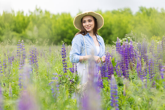 Beautiful young girl in a white dress, straw hat with a bouquet of violet flowers in her hands and picnic basket. Pretty woman in summer in the blooming field holding a bunch of purple lupin