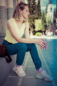 A young girl in a fashionable knitted T-shirt and dark jeans sits in the shadow of a building. Women's summer look.