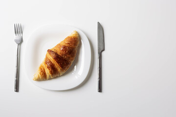 a freshly baked croissant lies on a white plate with a fork and a knife on the sides close-up isolated shot on a white...